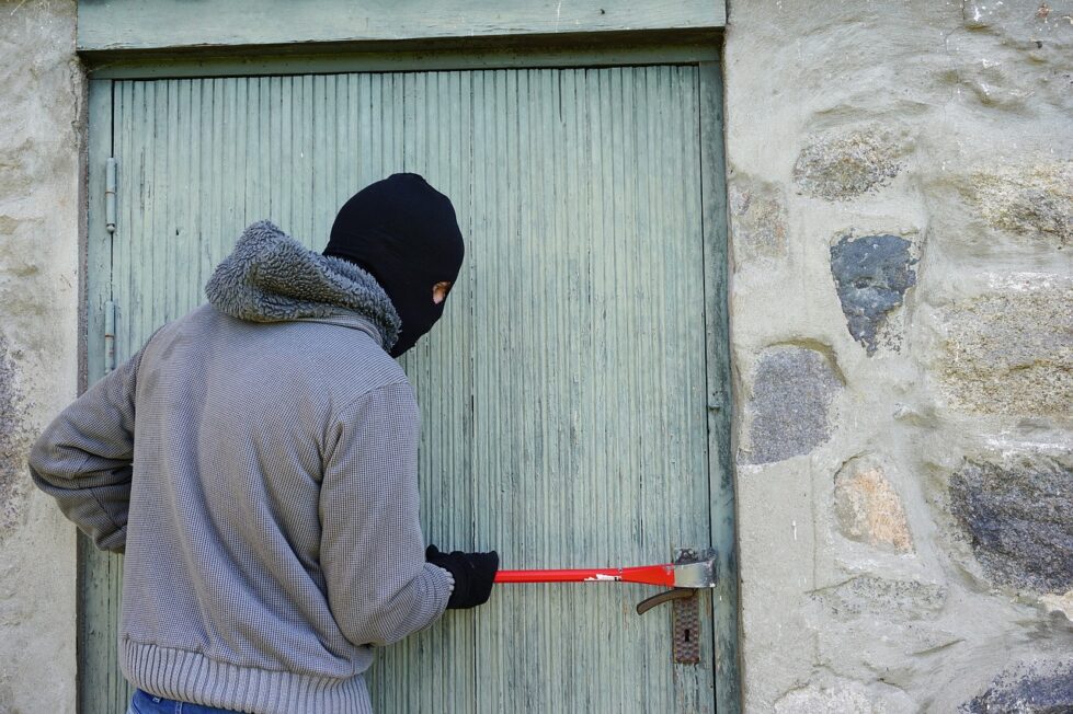 Masked burglar using a red crowbar to force open a wooden door